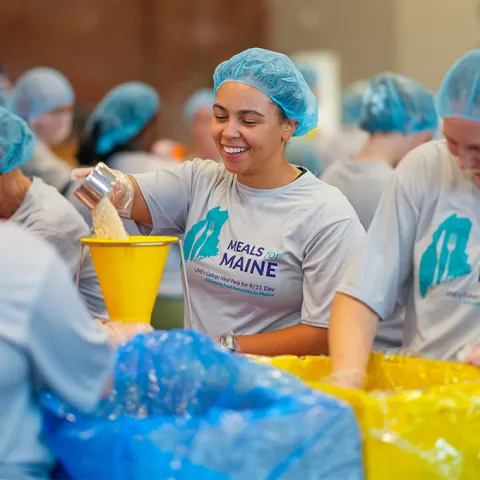 Volunteers in hairnets smile while packing bags of food at U N E's 2025 Meals for Maine event addressing food insecurity.